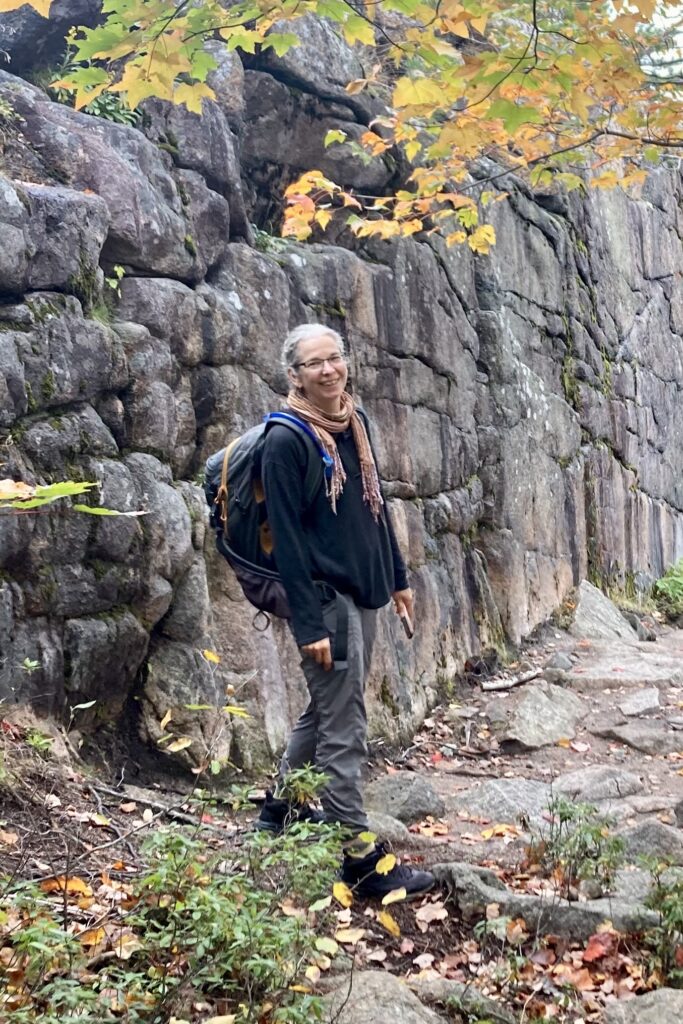 A person with a backpack stands on a rocky path, next to a large stone wall. They are surrounded by trees with autumn foliage in shades of green and orange. The ground is covered with fallen leaves.